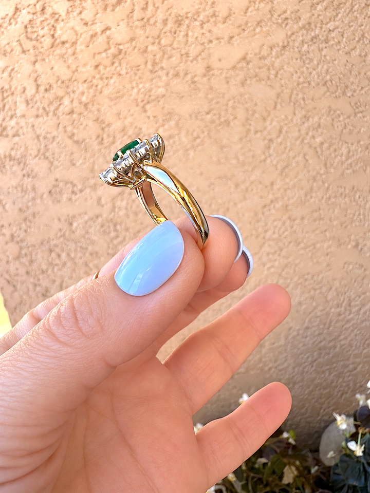 Bague marguerite émeraude et diamants or 18K tenue à la main, vue inclinée sur l'émeraude ovale, devant un mur beige