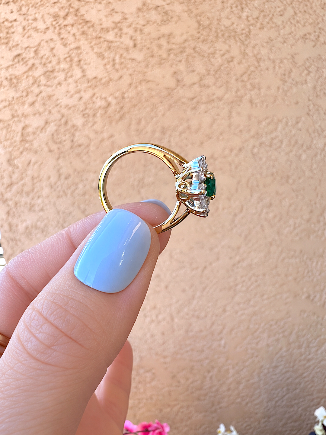 Bague marguerite émeraude et diamants or 18K tenue à la pince, vue de côté sur le jonc et l'émeraude ovale, devant un mur beige