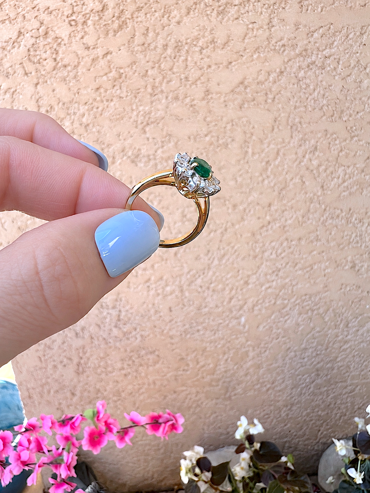 Bague marguerite émeraude et diamants or 18K tenue entre le pouce et l'index, vue de dessus sur l'émeraude, fleurs roses en arrière-plan