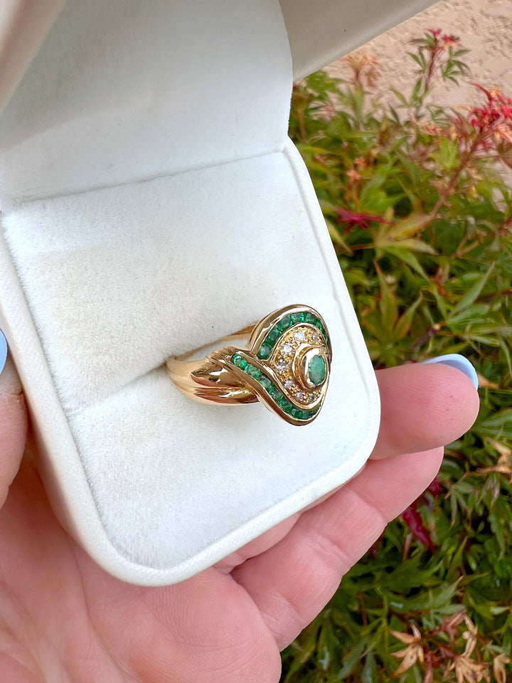 Bague émeraudes et diamants en or jaune 18K en écrin blanc tenu à la main, vue de côté sur fond de feuillage, joaillerie de seconde main