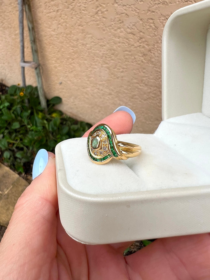 Bague en or jaune 18K ornée d'émeraudes et de diamants en écrin blanc, vue de trois quarts sur fond de mur beige, joaillerie de luxe d'occasion