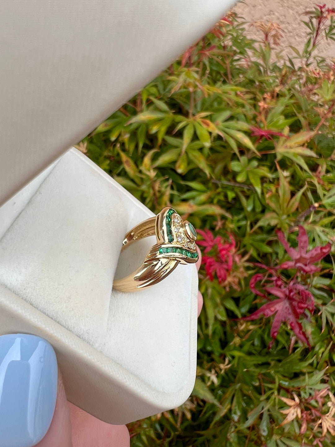 Bague de luxe de seconde main en or jaune 18K, émeraudes et diamants, vue de profil en écrin blanc sur fond de feuillage rouge