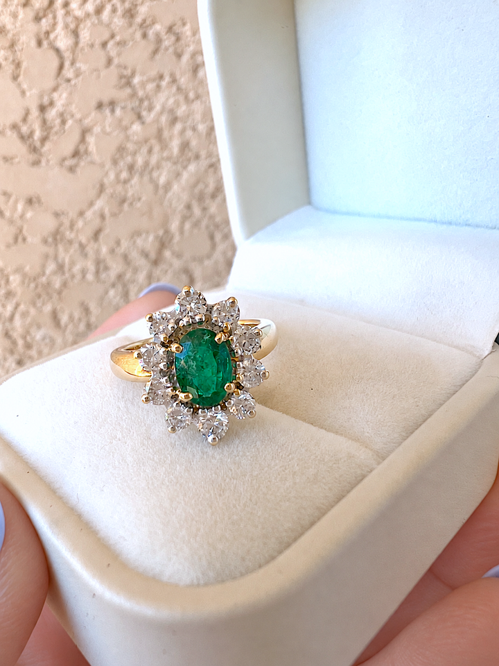 Bague marguerite émeraude et diamants or 18K dans un écrin ivoire tenu à la main, vue de trois quarts sur l'émeraude ovale et son entourage de diamants