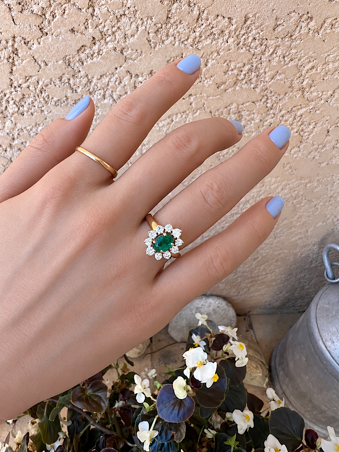 Bague marguerite émeraude et diamants or 18K portée à la main, ongles bleu pastel, avec un anneau fin en or sur le doigt voisin, devant des bégonias blancs