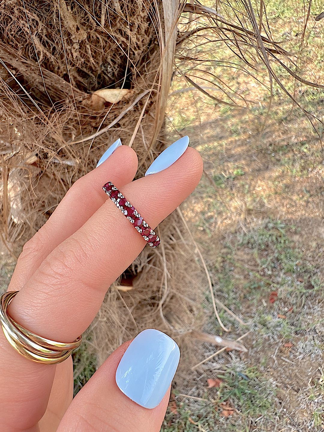 Alliance américaine rubis or gris 18K tenue entre deux doigts aux ongles bleu clair, devant un tronc de palmier