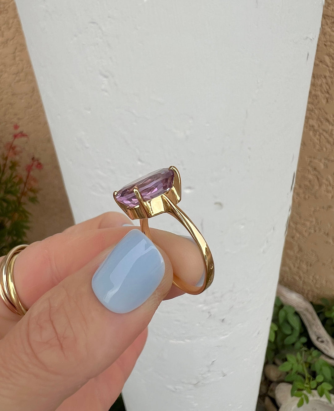 Bague solitaire améthyste or jaune 18K tenue entre deux doigts, vue de profil du jonc et de la monture, mur blanc
