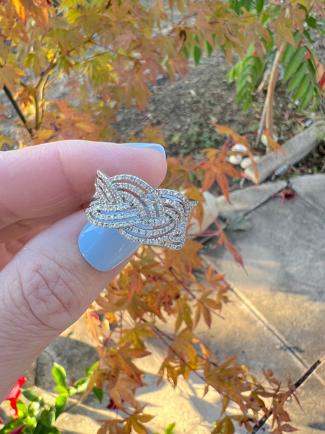 Bague tressée diamants or blanc 18 carats tenue entre les doigts, feuillage d'automne en arrière-plan