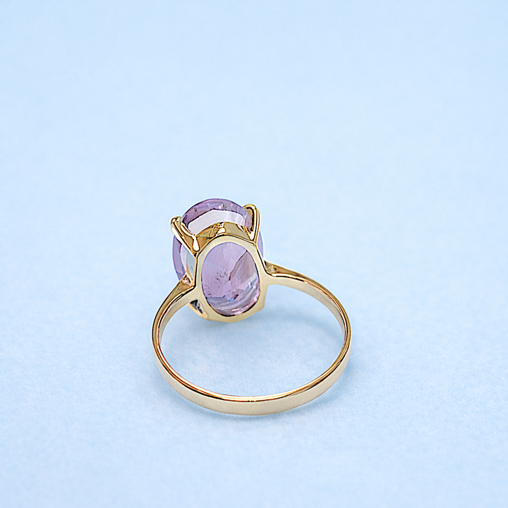 Bague en or jaune 18K avec pierre améthyste ovale en solitaire, monture griffe sur fond bleu.
