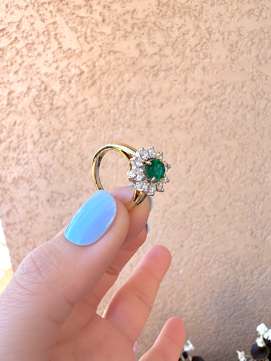 Bague marguerite émeraude et diamants tenue entre le pouce et l'index, vue de face sur l'émeraude ovale et le jonc en or jaune