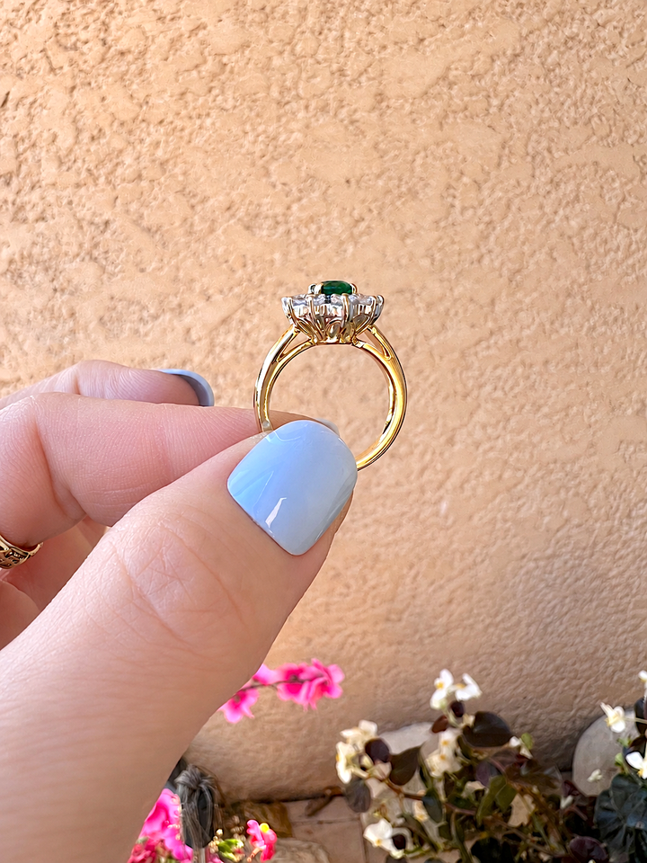 Bague marguerite émeraude et diamants or 18K tenue entre le pouce et l'index, vue du jonc fin en or jaune et de la corbeille de diamants
