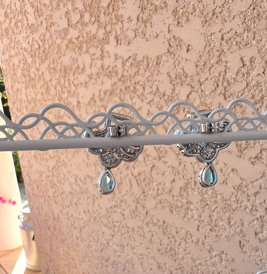 Dos des boucles d’oreilles or gris 750, topazes bleues et diamants, suspendues sur un support blanc, mur rose