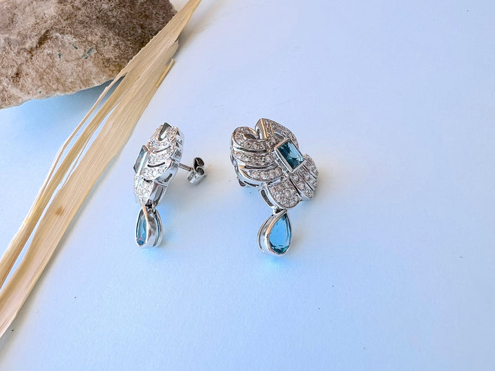 Boucles d’oreilles or gris 18K, topazes bleues et diamants, vue de côté sur fond bleu clair avec herbe séchée