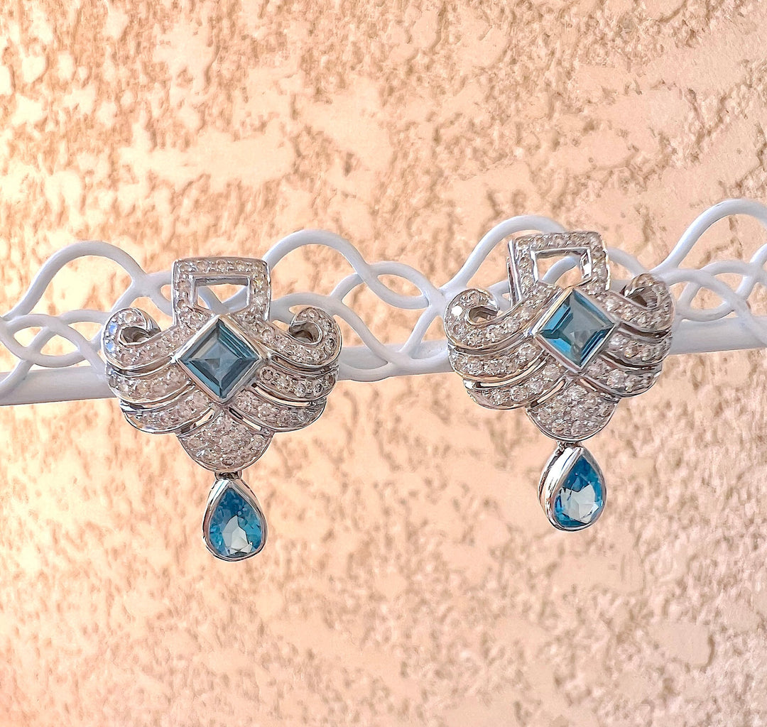 Boucles d’oreilles topazes bleues et diamants en or gris 18K, présentées sur un support blanc ajouré, fond rose