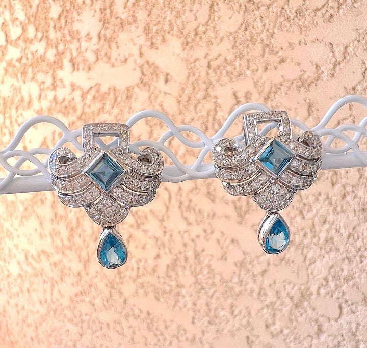 Boucles d’oreilles topazes bleues et diamants en or gris 18K, présentées sur un support blanc ajouré, fond rose