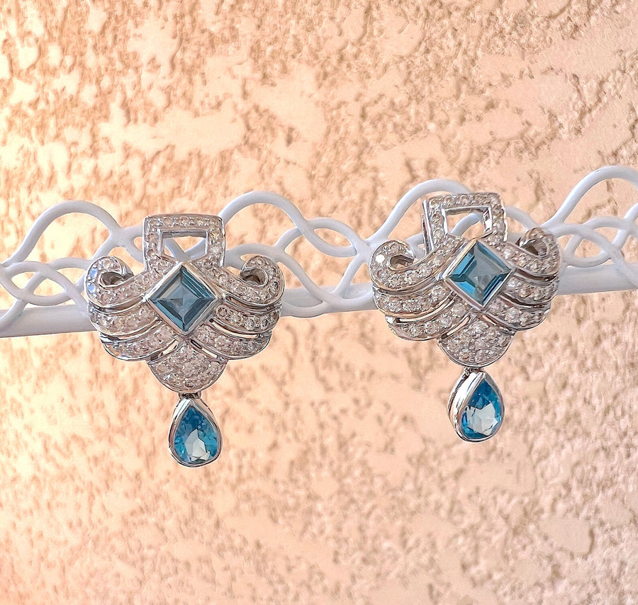 Boucles d’oreilles topazes bleues et diamants en or gris 18K, présentées sur un support blanc ajouré, fond rose