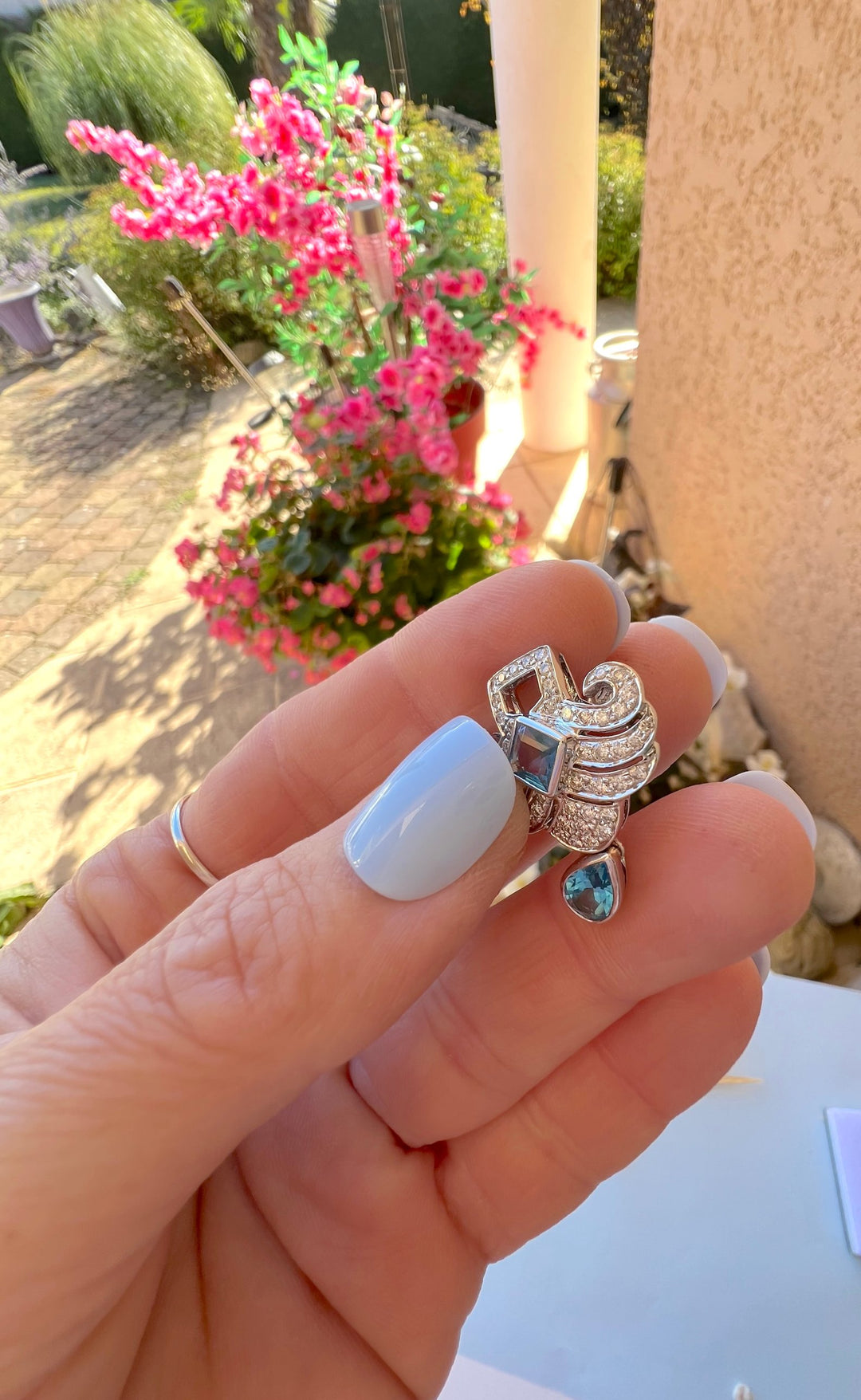 Boucle d’oreille topaze taille poire et diamants en or gris 18K tenue en main devant des fleurs roses