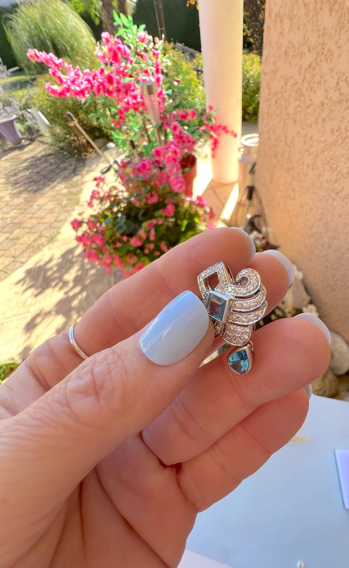 Boucle d’oreille topaze taille poire et diamants en or gris 18K tenue en main devant des fleurs roses