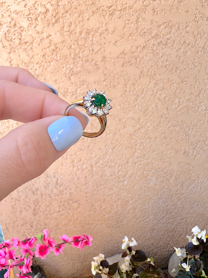 Bague marguerite émeraude et diamants or 18K tenue entre les doigts, ongles bleu clair, vue de trois quarts sur la pierre ovale, bégonias blancs en arrière-plan