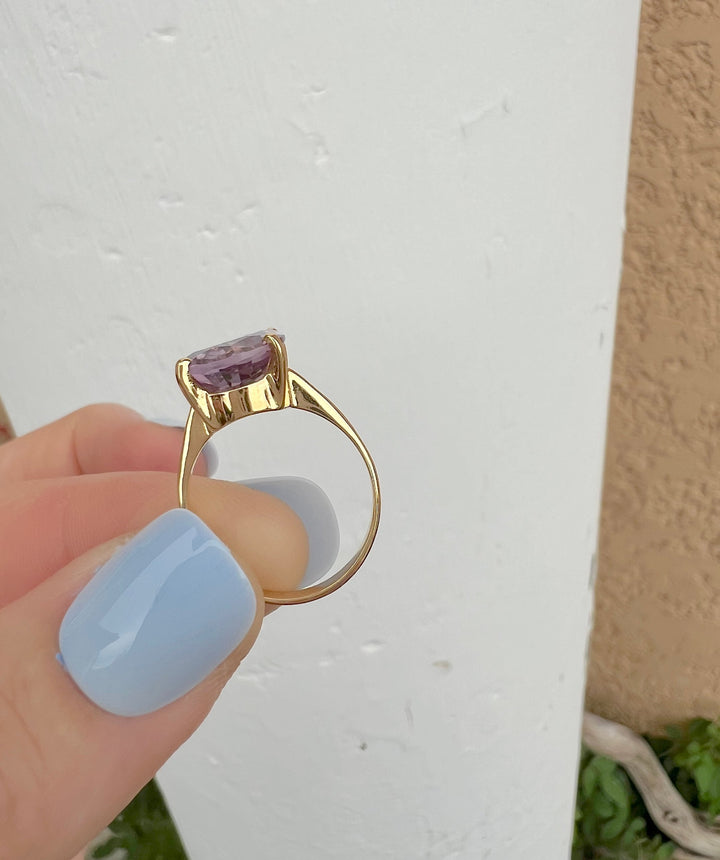 Bague solitaire améthyste ovale en or jaune 18K posée sur la tranche entre deux doigts, vue de profil, mur blanc