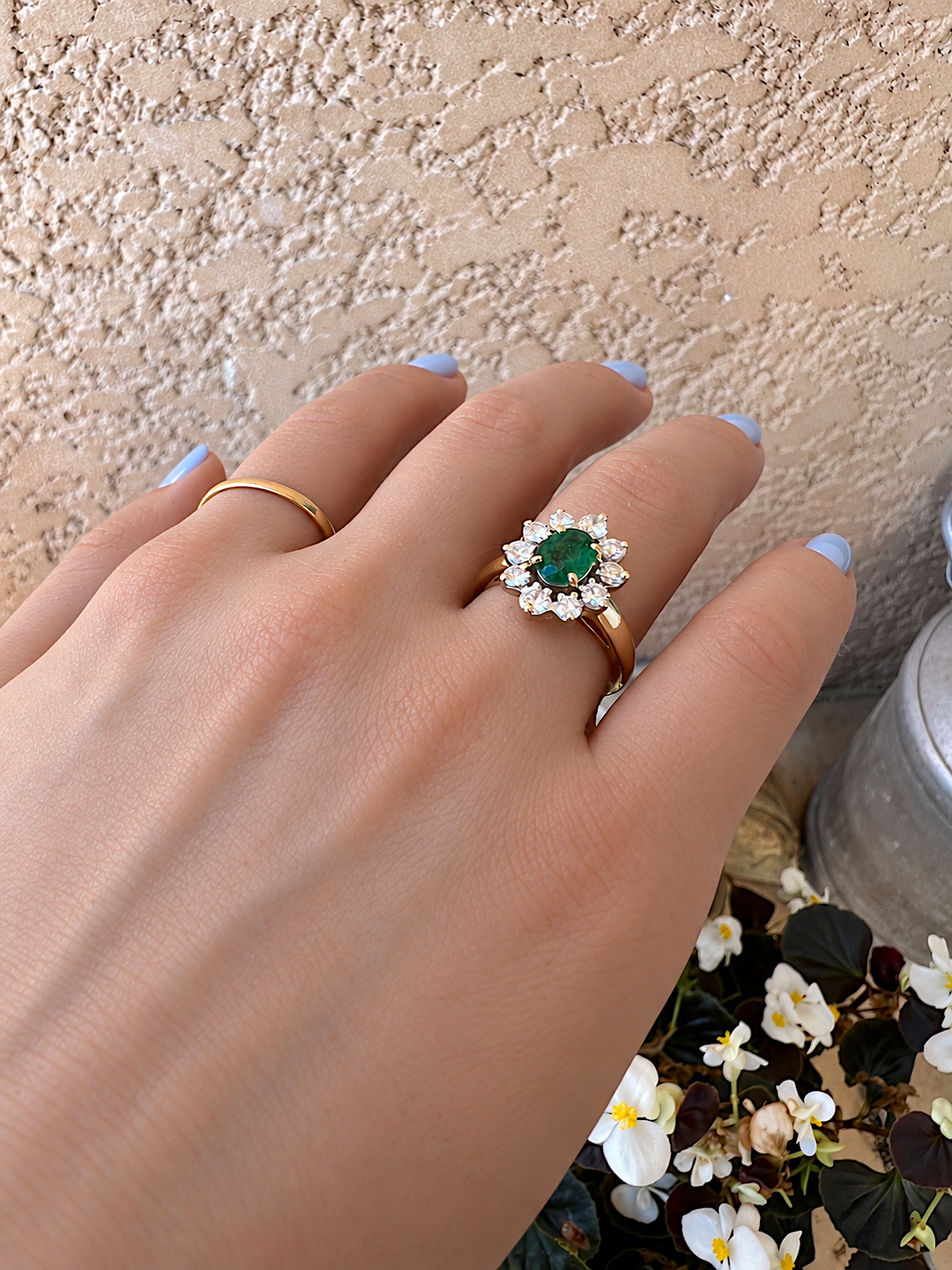 Main de femme portant la bague marguerite émeraude et diamants or 18K, ongles bleu clair, bégonias blancs et mur beige en arrière-plan