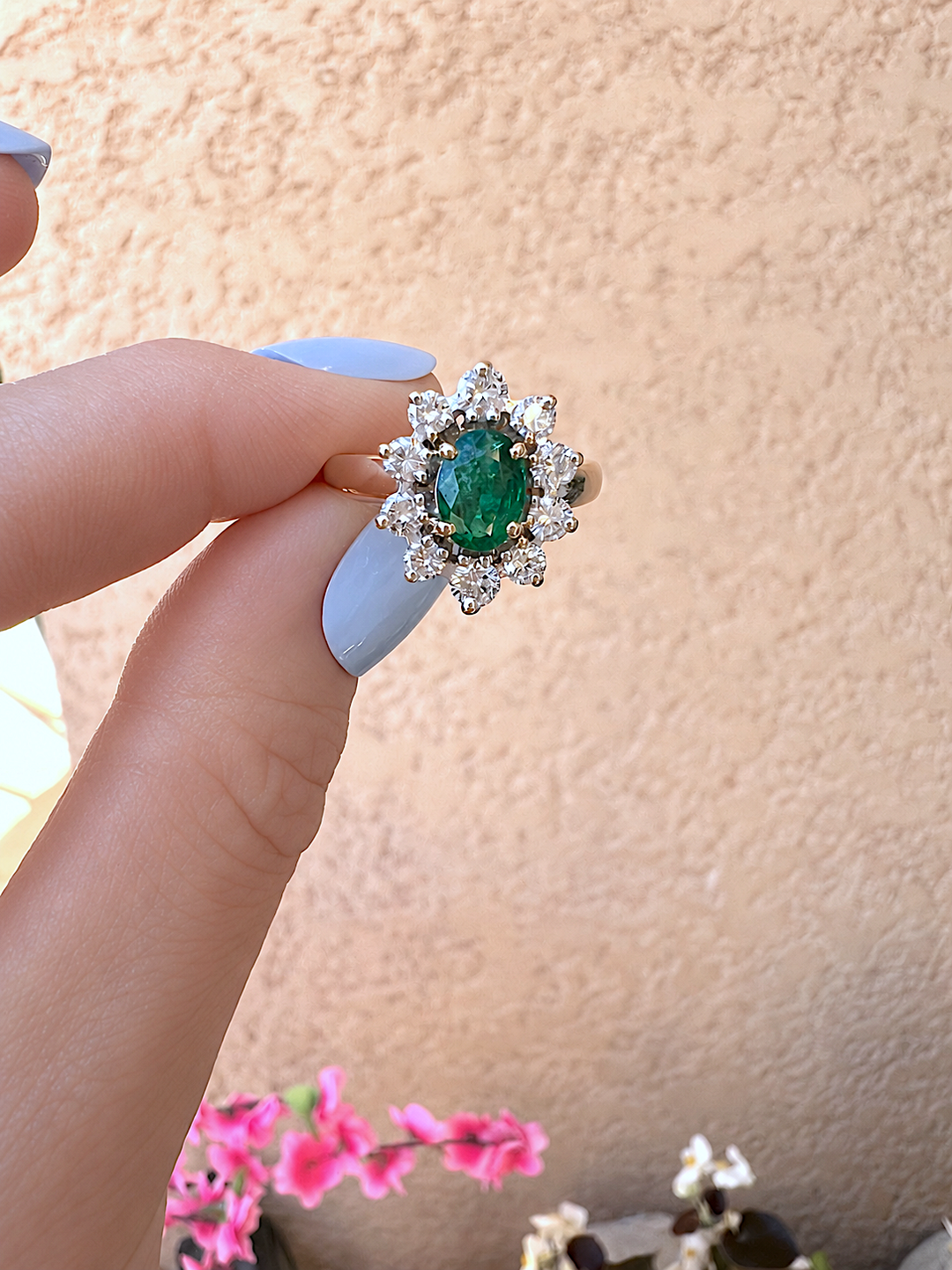 Bague marguerite émeraude et diamants or 18K tenue entre deux doigts, vue de face, ongles bleu clair, devant un mur beige
