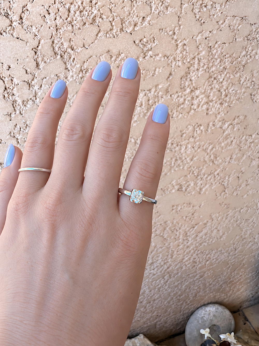 Main aux ongles bleu lavande portant le solitaire Chopard en platine 950 et une alliance fine en platine, sur mur beige